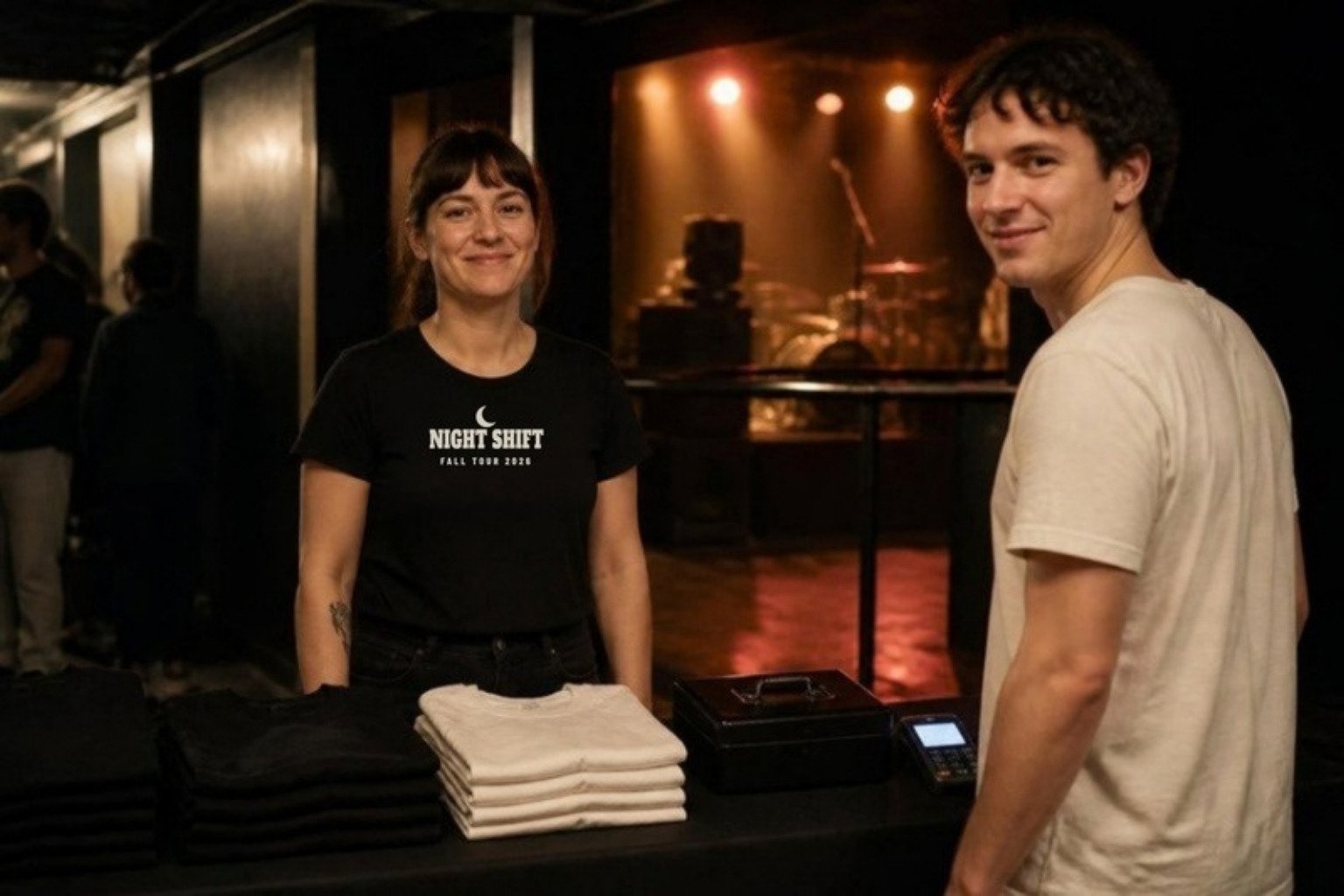 Demo: a bass player at the merch table of a small venue in a black tee printed with the fictional band name Night Shift, stacks of shirts on the table