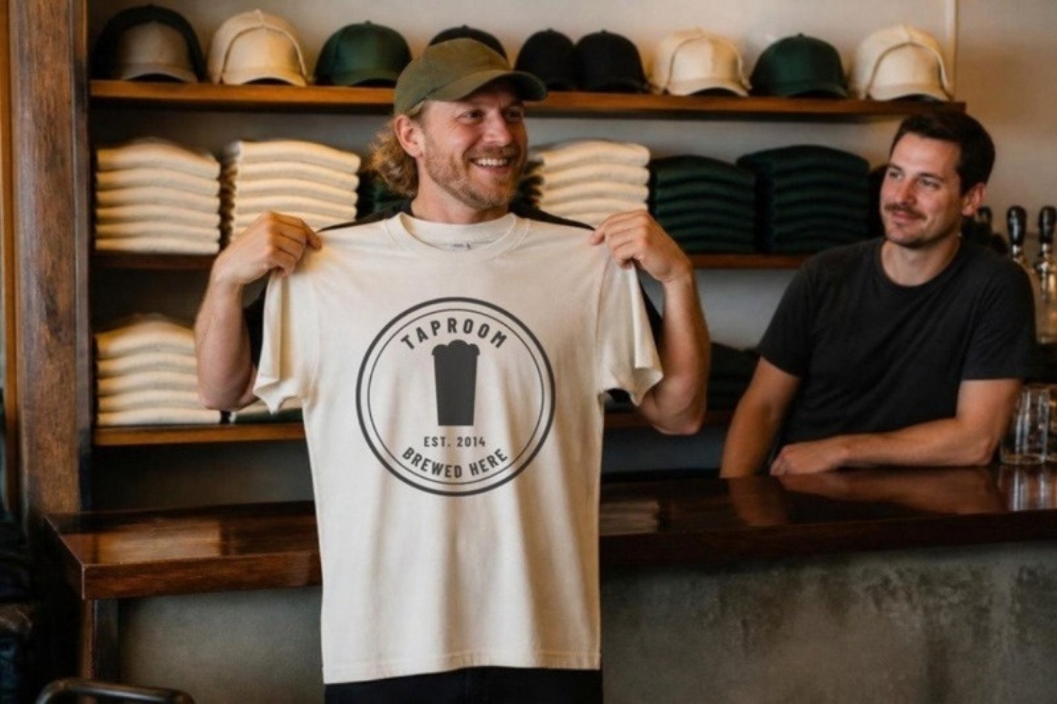 A guest at a taproom merch wall holds up a cream tee printed with the house badge, shelves of folded tees and plain caps behind