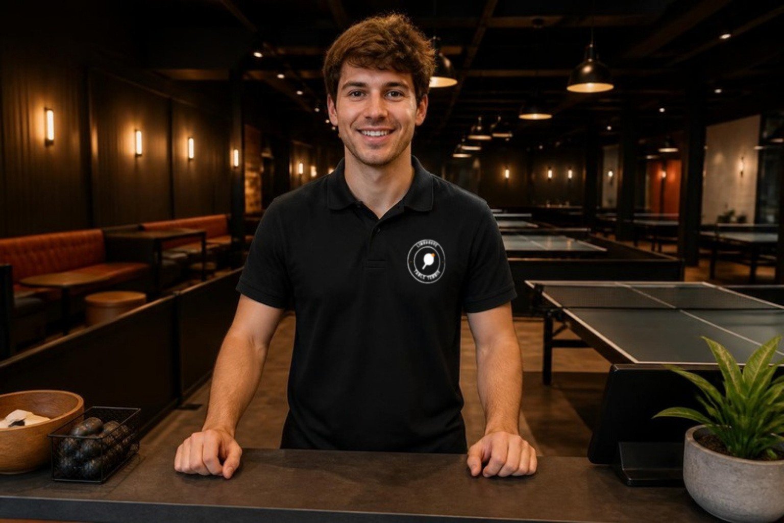 Demo staff member in a black polo with the Lindgrove crest at the front counter of a table tennis venue, tables behind